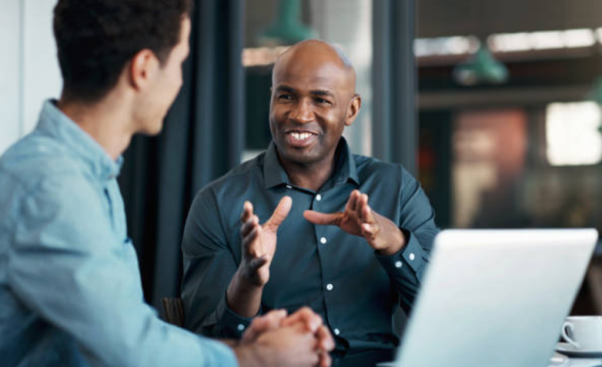 Professional man seated at table explaining a concept to a male colleague.